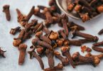 Whole dried cloves scattered on a pale surface beside a metal spoon